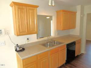 Kitchen with light wooden cabinets, beige countertops, and a double stainless steel sink. A black dishwasher is installed under the counter. A cordless phone is on the counter next to the wall. The floor is wooden.