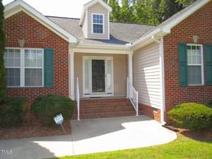 A brick house with a white trim and green shutters. The front door is recessed and flanked by two windows. A small porch with steps leads to the entrance, surrounded by neatly trimmed shrubs. A dormer window is visible above the door.