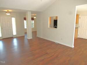 An empty living room with wood flooring and white walls. There's a doorway leading to another room, and a pass-through window above a counter. A ceiling light fixture is visible, and natural light comes in through a window near the door.