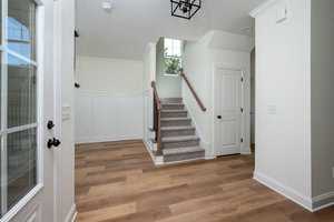 A bright entryway featuring wooden flooring and white walls. Stairs with a wooden banister lead upwards. A white door with a black handle is on the right, and a glass-paneled door is on the left. A ceiling light fixture hangs above.