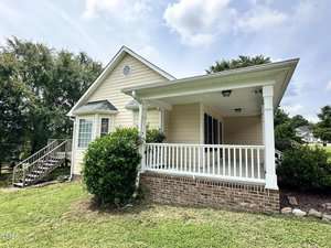 House with light-colored siding, a brick foundation, and a porch with white railings. Steps lead to the porch, and trees are visible in the background. The yard is grassy and the sky is partly cloudy.