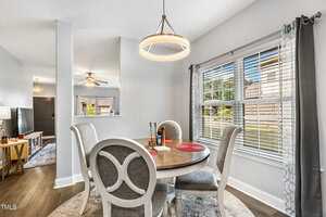 A cozy dining area with a round table and four upholstered chairs. A modern circular pendant light hangs above. A large window with blinds provides natural light. The room leads to a hallway with a view of a living area and ceiling fan.