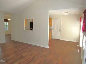 A spacious, empty room with light-colored walls and a vaulted ceiling. The wooden floor leads to a small room with a door and window on the right. A pass-through window from the kitchen is visible on the left. Natural light brightens the space.