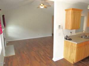 A bright living space with wooden floors and cream walls. A ceiling fan is above, and red curtains hang by a window. The open layout leads to a kitchen area with light brown cabinets and a double sink.