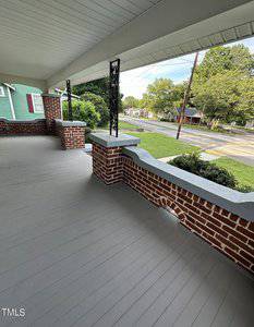 A porch with a covered roof and brick columns overlooks a quiet street lined with trees. The gray wooden floor complements the red brick walls, while the view includes a few houses and a lush green lawn.