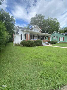 A white house with a gabled roof and brick foundation sits on a well-maintained lawn. A porch with columns extends from the front. There are shrubs near the porch, and a sidewalk leads to the entrance. A green house is visible nearby.