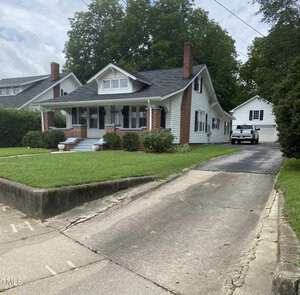 Nestled at 1103 College Street, this quaint American Craftsman-style house in Oxford features a wide front porch with brick pillars. The well-kept lawn and concrete driveway lead to a detached garage, framed by mature trees against a cloudy sky backdrop.