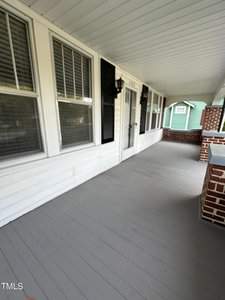 A white porch with a wooden floor, flanked by a row of windows and a door with black shutters. Two lantern-style lights are mounted beside the door. A brick railing with a green house visible in the background.