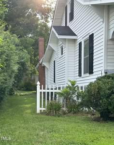 Side view of a white house with black shutters and a small white picket fence. Green shrubs and grass line the side, with sunlight peeking through the trees in the background.