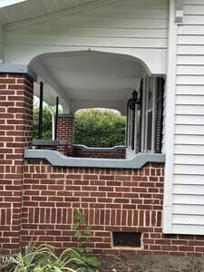 A house porch with a brick exterior and white siding. The porch has an arched opening, white railing, and a black lantern-style light fixture. Green shrubs are visible in the background.