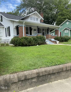 A charming white house with a brick chimney and a covered porch, surrounded by green bushes and a neatly trimmed lawn. A small set of stairs leads up to the porch. Tall trees are in the background.