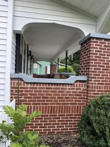 A brick porch with white wooden railings and a decorative brick pillar. The porch features a white overhang and black metal supports. A green house is visible in the background, and there are shrubs in the foreground.