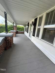 A spacious front porch with a grey wooden floor, white siding, and brick accents. Black shutters and multiple windows line the house wall, while a black light fixture hangs near the door. Lush greenery is visible beyond the porch railing.