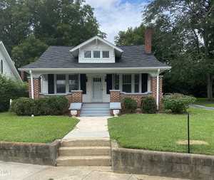 A small brick house with a gray roof and white accents, featuring a covered porch with white columns. It's surrounded by a green lawn and bushes, with a concrete path leading up to the entrance. The sky is partly cloudy.