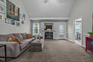 A spacious living room with a vaulted ceiling. It features a gray sectional sofa, a fireplace with a TV above, and a wall displaying family photos. A door leads to an outdoor area, and there are large windows with blue curtains letting in natural light.