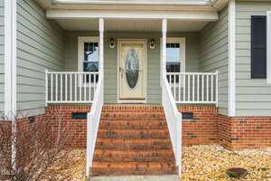 The image shows the front entrance of a house with green siding. It features a brick staircase leading to a wooden door with an oval glass panel. White railings and two windows flank the door. Sparse landscaping with stones and bare bushes is visible.