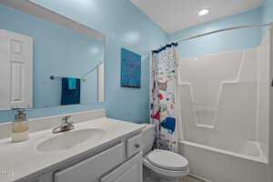 A clean bathroom with light blue walls features a white vanity with a sink and a mounted mirror above. A decorative picture hangs on the wall. A shower with a colorful curtain is next to a toilet.