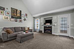 A cozy living room with a green sectional sofa and ottoman, decorated wall with family photos and artwork, a fireplace with a TV above it, and a door with a curtain. The carpet is brown, and natural light fills the room.