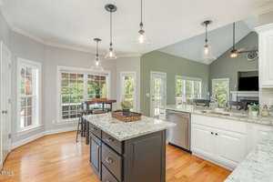Bright kitchen with hardwood floors, featuring an island with a dark base and granite countertops. Pendant lights hang from the ceiling. Large windows provide natural light, and there's a door leading to an outdoor area. White cabinetry lines the walls.