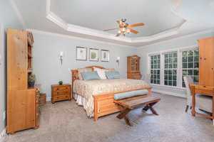 A spacious, elegantly designed bedroom with a wooden bed, matching nightstands, and a bench. The room features a tray ceiling with a ceiling fan, a large window letting in natural light, and neutral-toned decor for a relaxing atmosphere.