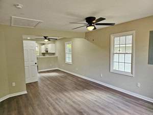A bright, empty room with light wood flooring, beige walls, and large windows. Ceiling fans with lights are present. A door leads to a kitchen area with white cabinets and a small counter. Natural light fills the space.