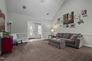 A cozy living room with a high ceiling, featuring a gray sectional sofa and coffee table. The walls are adorned with various photo frames and artwork. The room is well-lit by a floor lamp beside the sofa, and a doorway leads to a sunlit area.