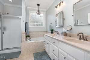 A modern bathroom with a shower, bathtub, and a double sink vanity. The room features white cabinets, a large mirror, and two light fixtures. A small window lets in natural light, and a gray towel hangs near the bathtub.