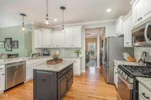 Modern kitchen with white cabinets, stainless steel appliances, and a central island with granite countertops. Pendant lights hang above the island. Hardwood floors complement the light green wall and decor accents.