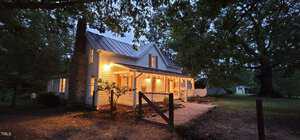 A charming white house with a pitched roof and front porch, warmly lit in the evening. Surrounded by large trees, a fence outlines the yard with a gravel path leading to the entrance.