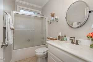 A small, well-lit bathroom with a frosted glass shower, white toilet, and a vanity with a round mirror. The sink area has a tissue box, basket of towels, and a vase of flowers. A towel hangs on a chrome bar by the door.