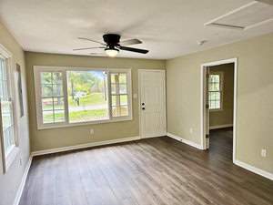 A bright, empty room with beige walls and wooden flooring. It features a ceiling fan, large windows with a view of greenery outside, and a white door leading to another room. Natural light illuminates the space.