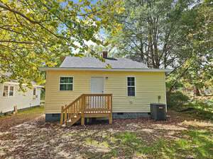 A small yellow house with a simple porch and a single door, surrounded by trees and grass. An outdoor air conditioning unit is visible on the side. The sky is clear and sunny.