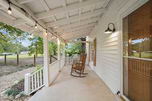 Front porch of a white house with a wooden rocking chair and string lights hanging from the ceiling. The porch overlooks a spacious yard with trees and a gravel path.