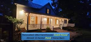 Nestled in Franklinton, this charming white farmhouse at 1676 Suitts Store Road boasts a wraparound porch illuminated by string lights at dusk. The renovated 1862 farmhouse/guest house sits on almost 13 acres, featuring a tranquil pond and a horse barn.