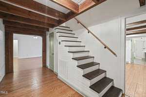 A hallway with wooden floors and exposed ceiling beams, featuring a white staircase with dark wood steps. Two doorways lead to adjoining rooms, one visible with white walls and wooden flooring.