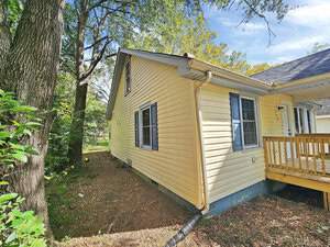 A yellow house with a sloped roof and blue-trimmed windows, surrounded by trees and greenery. There is a wooden porch on the side and a downspout for drainage. The sky is clear and blue.