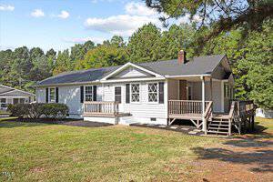 A single-story house with light gray siding, a dark roof, and a wooden front porch. It is surrounded by a grassy yard and tall trees in the background under a partly cloudy sky.