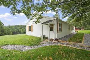 A small white house with a sloped roof sits on a grassy yard, surrounded by trees. The house has wood-framed windows and a gravel path leading to the door. A pond is visible in the background under a partly cloudy sky.