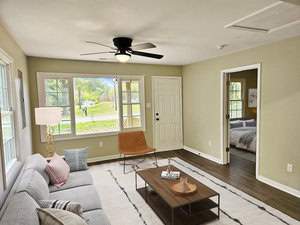 Modern living room with a light gray sofa, pillows, and a coffee table on a white rug. A brown chair and a standing lamp are in front of large windows. A ceiling fan is above, and an open door leads to a bedroom with a neatly made bed.