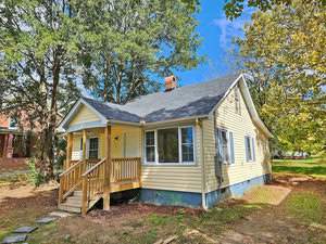 A small, single-story yellow house with a front porch and steps, surrounded by tall trees under a bright blue sky. The yard is spacious with scattered leaves, and the house has several windows visible from the front.