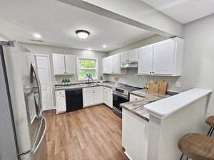 A modern kitchen with white cabinets, stainless steel appliances, and wood flooring. It has a double-door fridge, dishwasher, oven, and black sink. Natural light comes through a window above the sink, and there are two wooden bar stools at a counter.