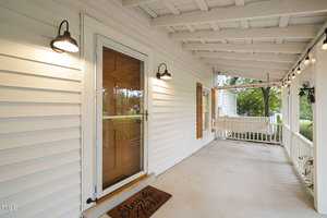 A white wooden porch with a doormat that reads "Stay Awhile." The porch features a glass storm door, two wall-mounted lights, string lights along the ceiling, and a distant view of a swing and greenery.