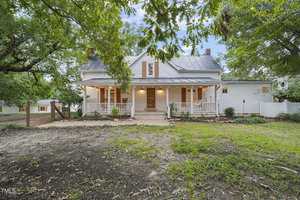 A charming white farmhouse with a large front porch, surrounded by lush greenery. The house features a gabled roof, brick walkway, and white picket fence, creating a picturesque rural setting under a clear sky.