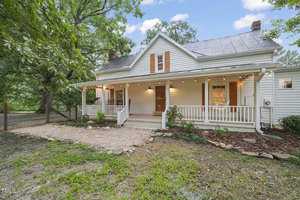 A charming white house with a wrap-around porch adorned with string lights. Surrounded by lush greenery and trees, the house features a gravel path leading to the entrance and wooden rocking chairs on the porch.