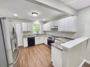 Modern kitchen with white cabinets, stainless steel appliances, and a wood laminate floor. The room features a double sink under a window, a black dishwasher, and an oven with a range hood. A partial wall separates the kitchen from another area.