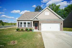 A single-story house with beige siding and a large white garage door. The front yard features a neatly manicured lawn and a small garden. A clear blue sky with scattered clouds serves as the backdrop.