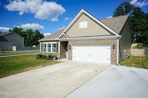 A modern single-story house with a brick facade and beige siding, featuring a two-car garage and a well-maintained front yard. A concrete driveway leads to the garage, and the sky above is partly cloudy.