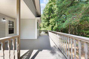 A spacious, wooden deck attached to a house, lined with a railing, extends towards a view of lush green trees on a sunny day.