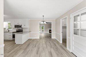 A spacious, modern kitchen and dining area with light wood flooring. The kitchen features white cabinets, granite countertops, and stainless steel appliances. A chandelier hangs above the dining area, and a fireplace is visible in an adjacent room.