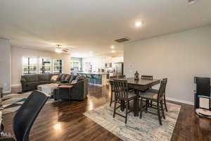 Open-plan living room with dark wood flooring, featuring a large gray sectional sofa, black dining table with chairs, and a kitchen with white cabinets and stainless steel appliances. Large windows in the background offer natural light.